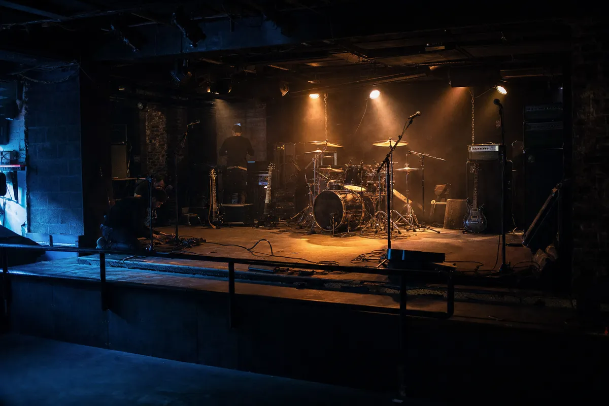 Palco di un piccolo club con batteria, amplificatori, microfoni e chitarre durante il soundcheck, luci calde e fumo