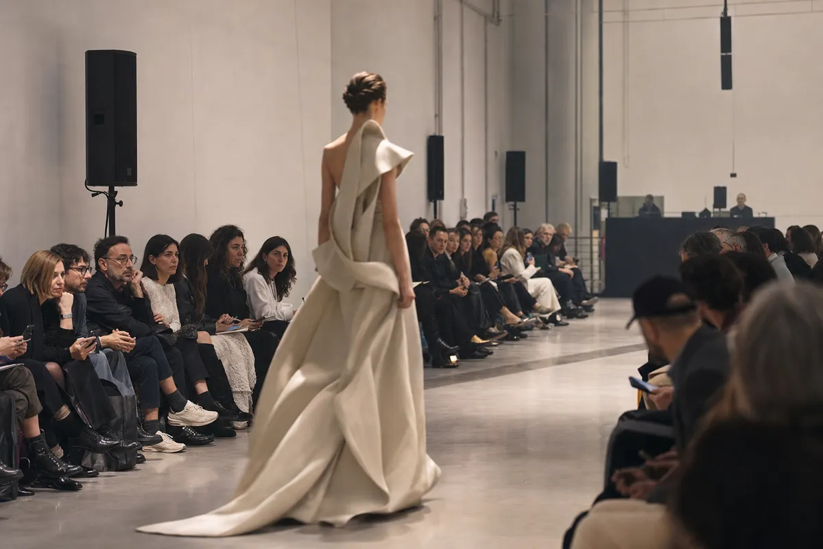 Modella in abito haute couture color avorio sfila su passerella minimale davanti a pubblico seduto e casse audio
