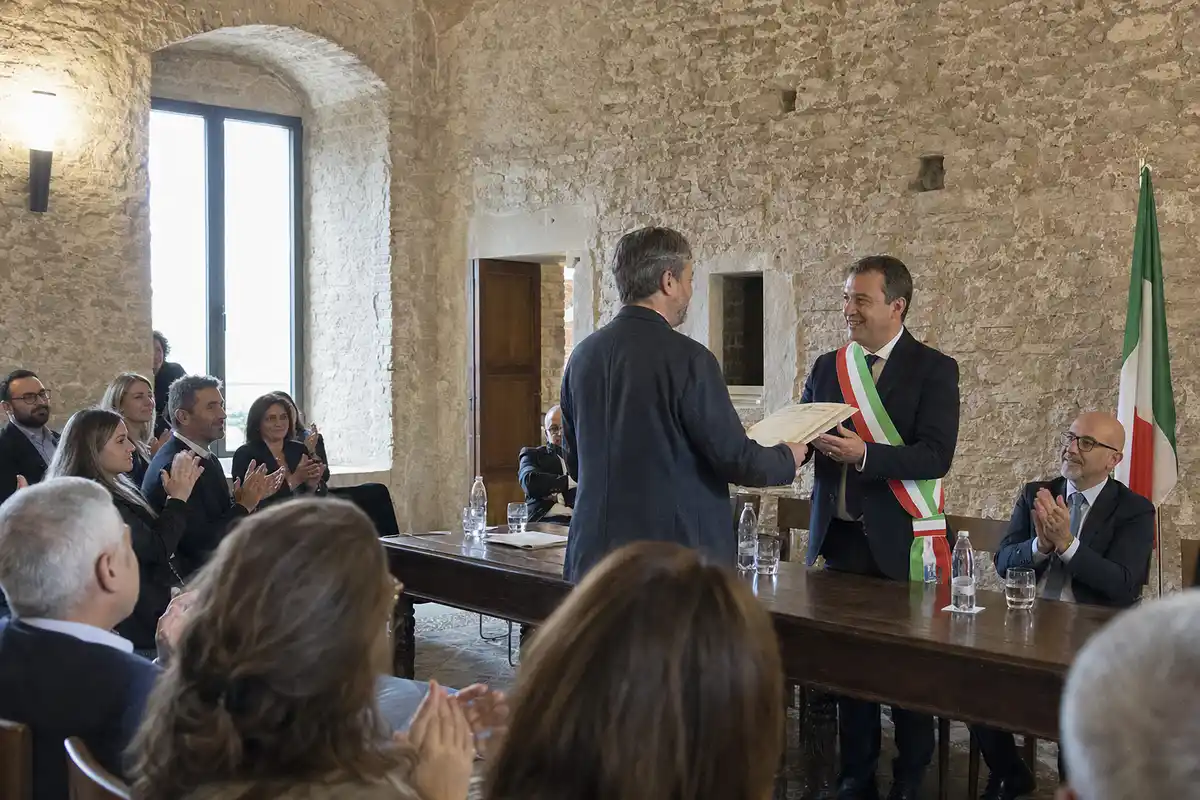 Cerimonia comunale in sala in pietra: sindaco con fascia tricolore consegna un attestato a un uomo, pubblico applaude