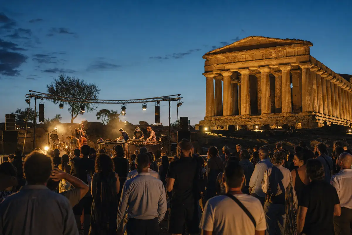 Pubblico davanti a un palco serale con luci calde, con un tempio greco illuminato sullo sfondo al crepuscolo