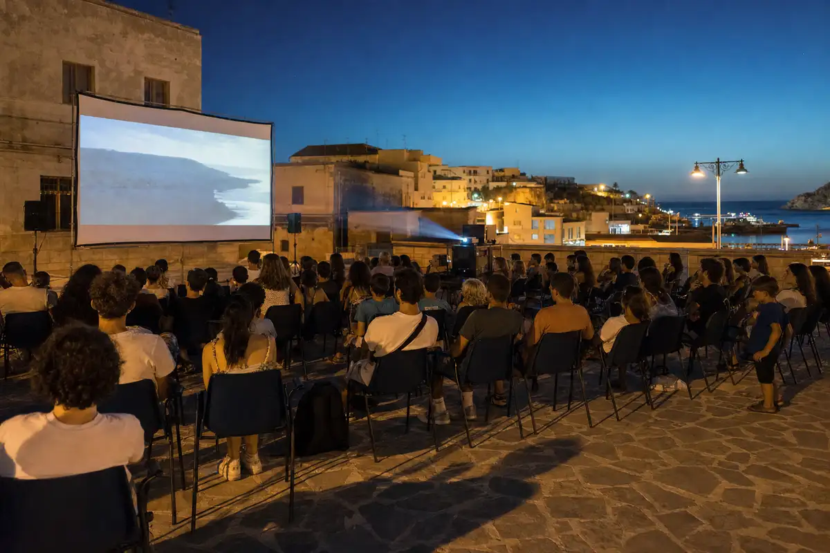 Pubblico seduto a una proiezione di cinema all’aperto al tramonto, con schermo e fascio del proiettore sullo sfondo