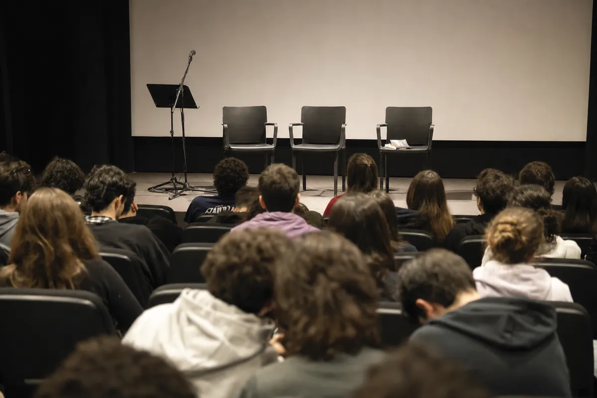 Pubblico giovane seduto in sala cinema davanti a palco con microfono, tre sedie vuote e grande schermo bianco
