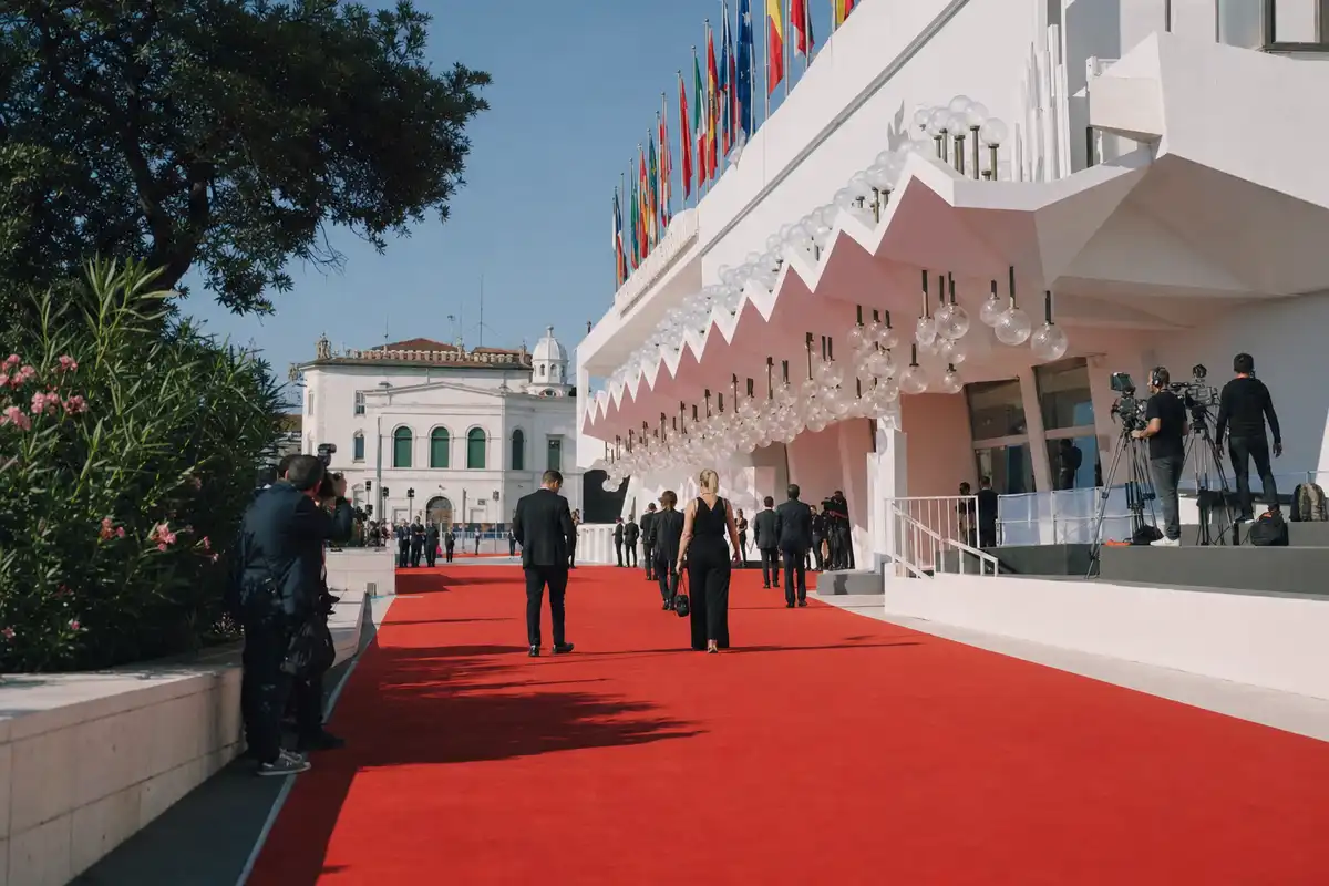 Red carpet davanti al Palazzo del Cinema al Lido di Venezia, con bandiere, luci e fotografi sullo sfondo