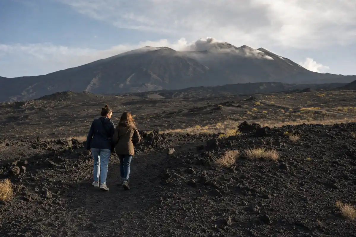 Due donne camminano su un sentiero di lava scura verso l’Etna con un pennacchio di fumo sotto un cielo nuvoloso