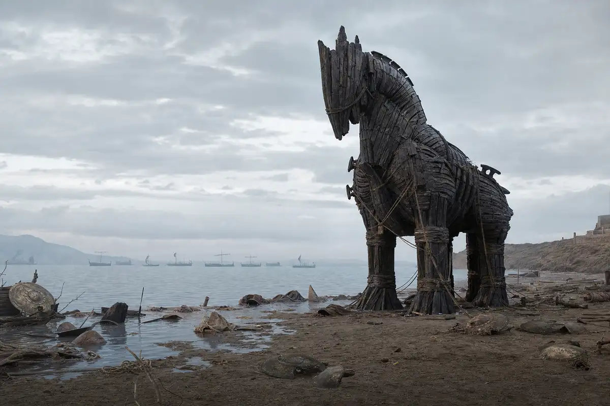 Cavallo di legno abbandonato sulla spiaggia, con scudi rotti e navi all’orizzonte sotto un cielo grigio