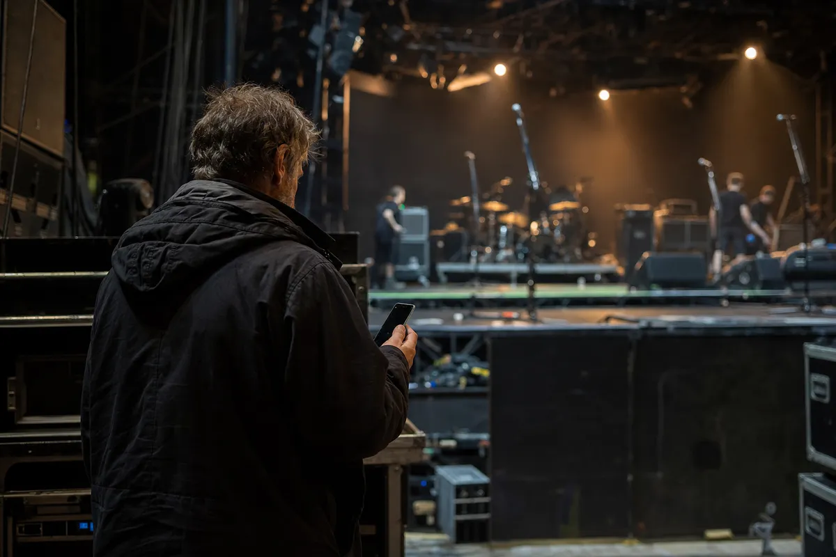 Uomo di spalle con smartphone davanti a un palco in allestimento, con luci calde, microfoni e batteria