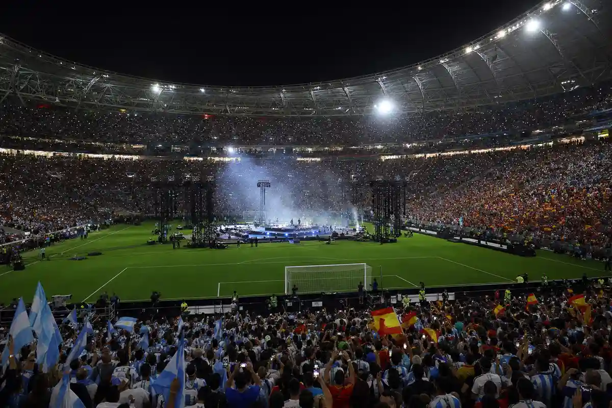 Stadio pieno di notte con palco e luci al centro del campo durante l’intervallo, tifosi con bandiere di Spagna e Argentina