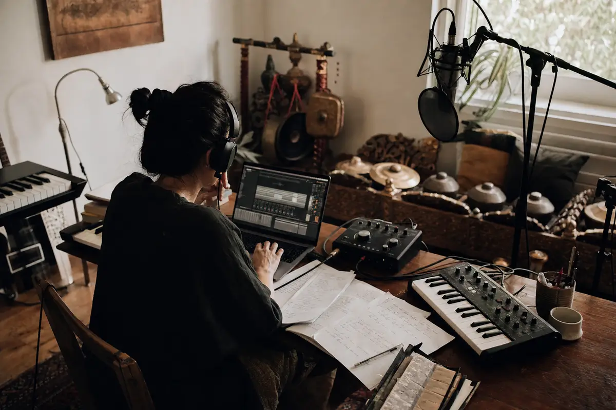Musicista di spalle in home studio con laptop, tastiera MIDI, microfono e appunti, con piccoli gong sullo sfondo