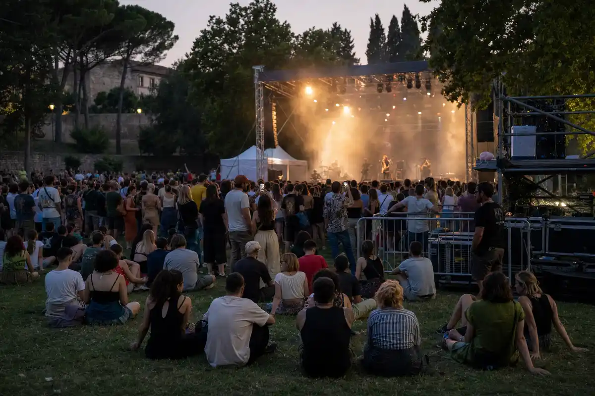 Pubblico seduto e in piedi su un prato davanti a un palco illuminato al tramonto, con luci calde e foschia