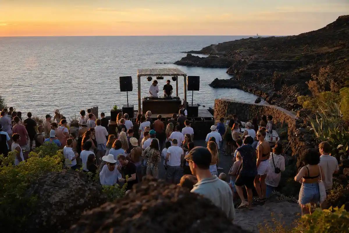 Piccolo palco con dj e folla al tramonto su una costa rocciosa, con il mare sullo sfondo