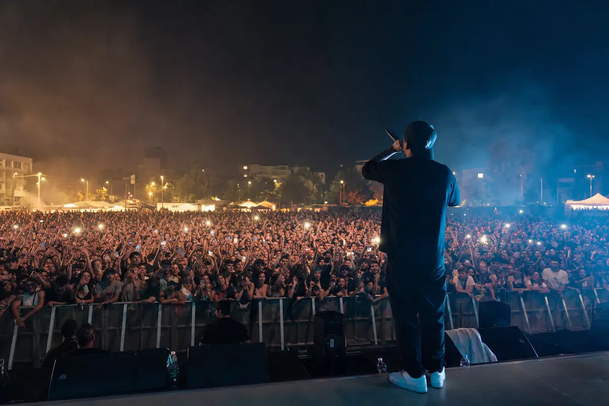 Rapper di spalle con microfono sul palco davanti a una folla in festival notturno con luci e smartphone alzati