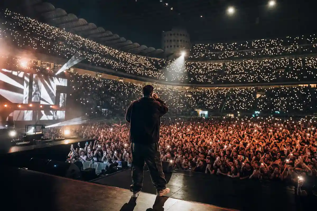 Rapper di spalle sul palco con microfono davanti a uno stadio pieno illuminato da migliaia di luci degli smartphone
