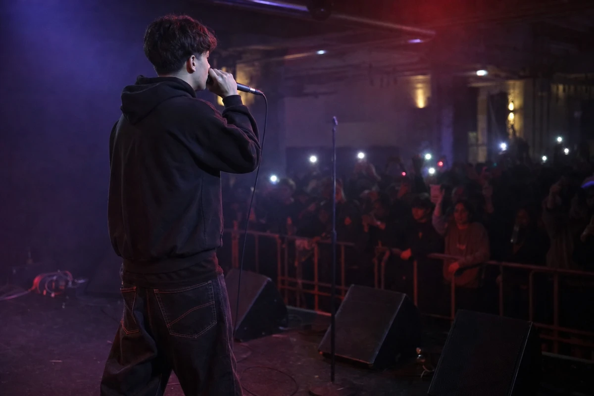 Rapper di spalle con microfono su un palco, davanti a un pubblico con luci di smartphone in un club