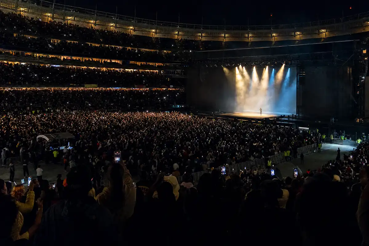 Concerto notturno in uno stadio gremito, con palco minimal illuminato e artista in silhouette tra luci e fumo